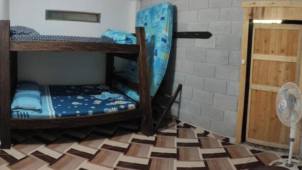 Una cama individual con ropa de cama azul, un ventilador y una puerta de madera en una habitación con suelo estampado.