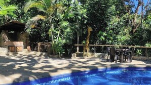 Zona de piscina amb sòl de rajola, envoltada de vegetació exuberant i una construcció de pedra.