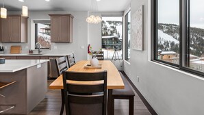 A modern kitchen with a central island, wooden dining table, and chairs. There is a large window providing a view of a snowy landscape. - Modern Mountainside Retreat | Hot Tub & Views (Sandpoint)
