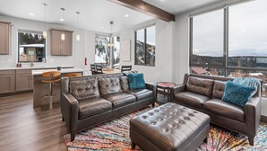 A spacious living room with a brown leather sofa and ottoman, a colorful rug, and large windows offering a view of a snowy landscape. - Modern Mountainside Retreat | Hot Tub & Views (Sandpoint)