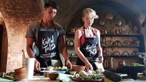 Dos personas con delantales preparando comida en una cocina rústica con estantes llenos de vajilla y una pared de piedra.