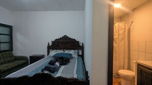 Ein kleiner, schlichter Raum mit einem Bett, einem Nachttisch, einem grünen Schlafsofa und einem Badezimmer mit Toilette und Dusche.