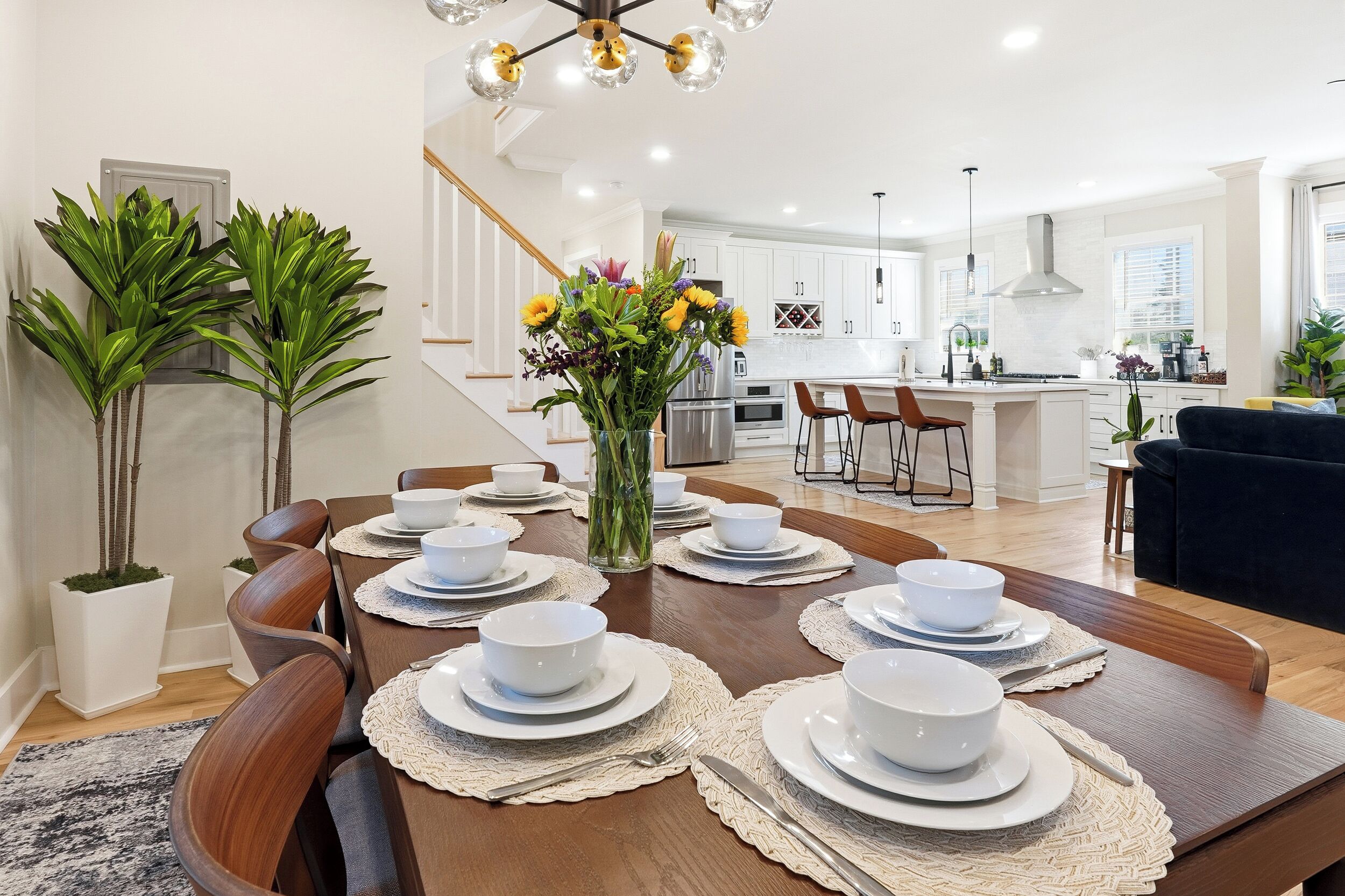 A modern dining area with a wooden table set for six, a vase with flowers, and a kitchen in the background.