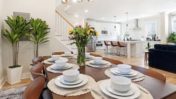 A modern dining area with a wooden table set for six, a vase with flowers, and a kitchen in the background.