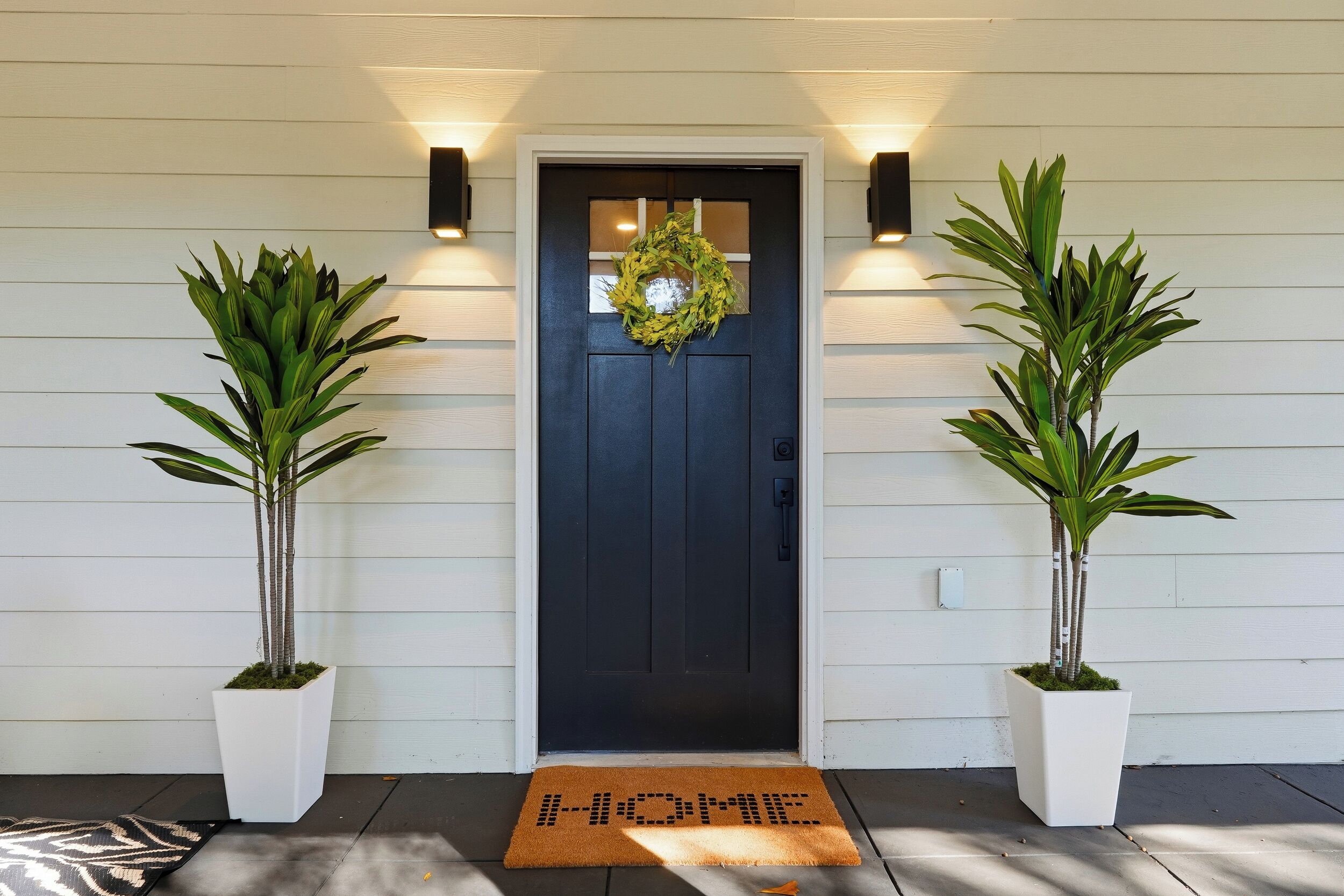 A black door with a wreath, flanked by two potted plants, and a doormat with "HOME" written on it.