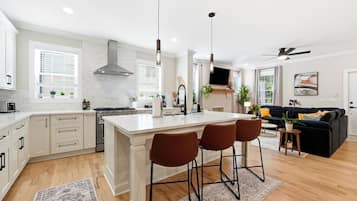 A modern kitchen with a central island, bar stools, and a dining area with a black sofa and yellow cushions.
