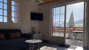 Een moderne woonkamer met een bank, een kleine tafel en een tv aan de muur bevestigd. Er is een grote schuifdeur die leidt naar een buitenruimte met een tafel en stoelen.