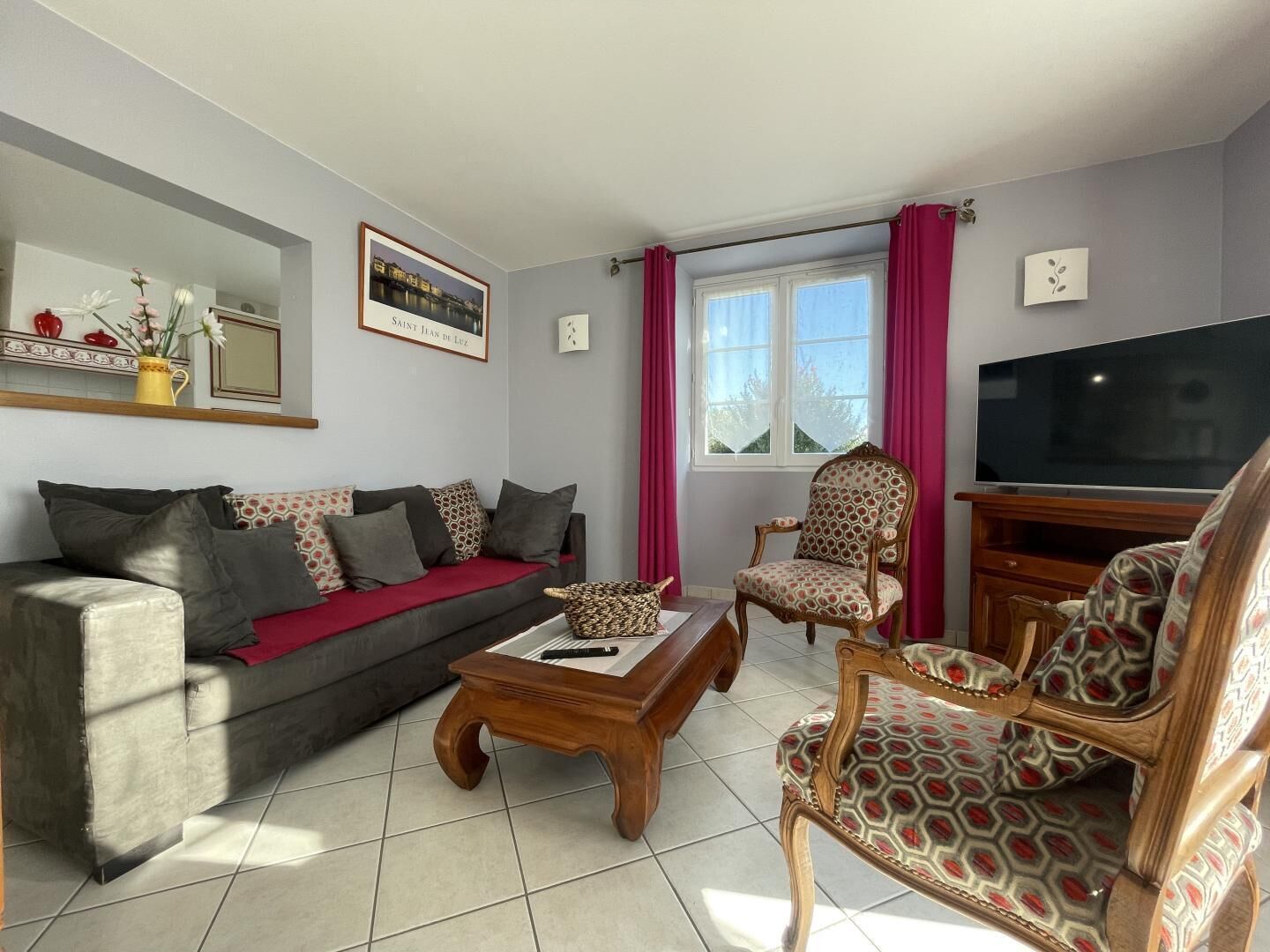 A living room with a sofa, a wooden coffee table, a patterned armchair, a television, and a window with curtains.