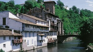 A historic town with buildings along a river, a stone bridge, and lush greenery in the background. - URTXOAK BIXTAN - UHART-CIZE (UHART-CIZE)