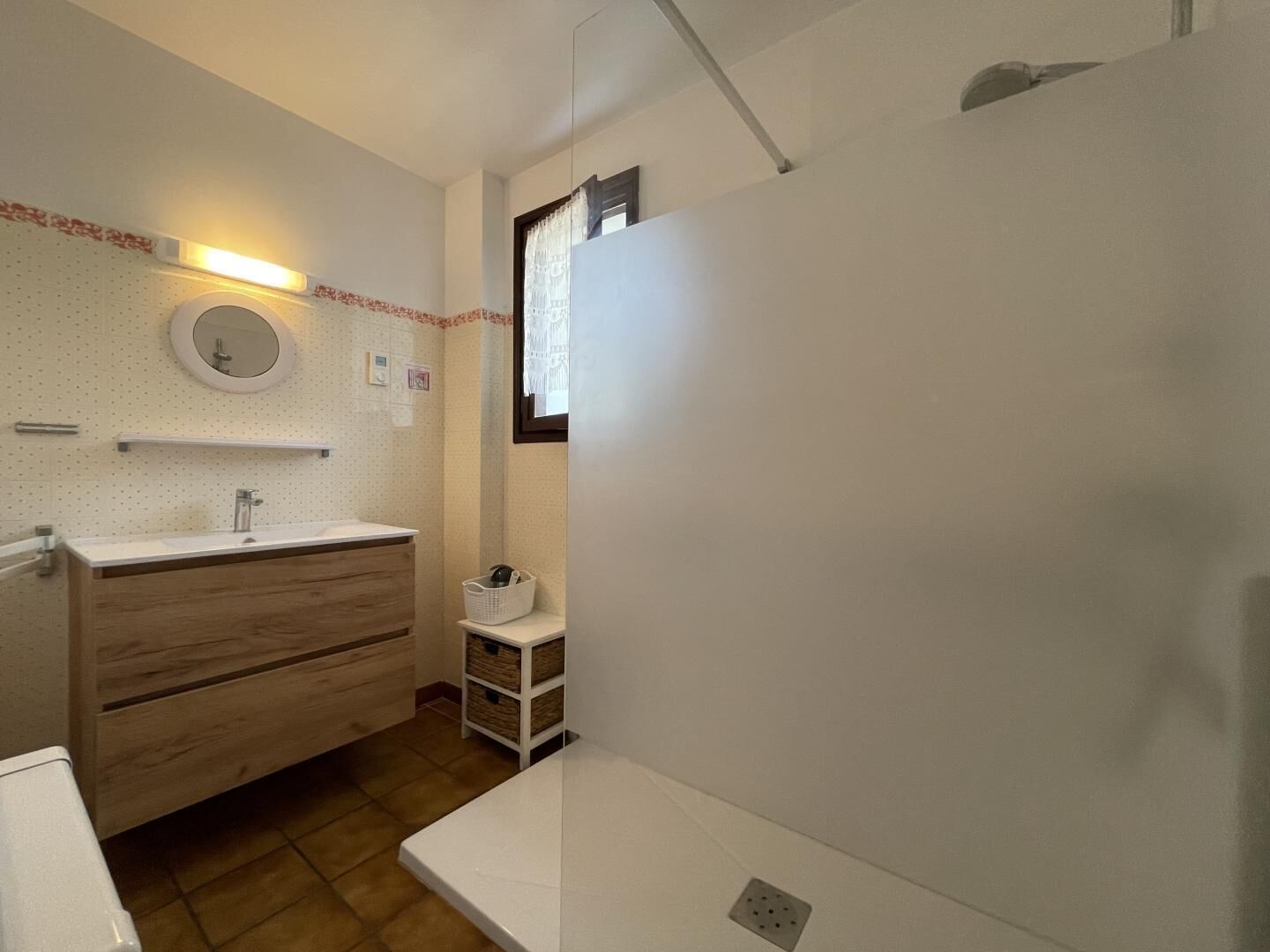 A bathroom with a wooden vanity, a round mirror, and a small window.