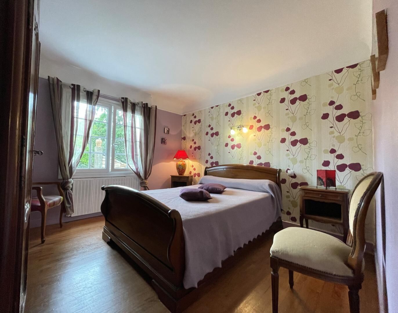 A bedroom with a wooden bed, a chair, a nightstand with a lamp, and a window with curtains.