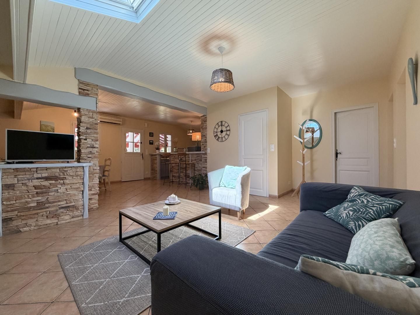 A spacious living area with a stone accent wall, a flat-screen TV, a dark sofa with patterned cushions, and a dining table with a candle.