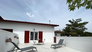Un patio con dos sillones de descanso y una mesa, un edificio blanco con contraventanas rojas, y una palmera al fondo.
