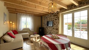 Una sala de estar acogedora con un sofá, una mesa de comedor con mantel rojo y blanco, una pared de piedra y un ventanal con vistas a la vegetación.