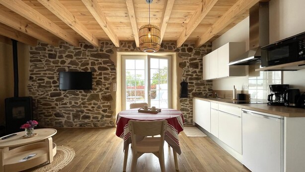 Una habitación acogedora con pared de piedra, techo de madera, una mesa de comedor con sillas, una cocina con armarios blancos y un televisor instalado en la pared.