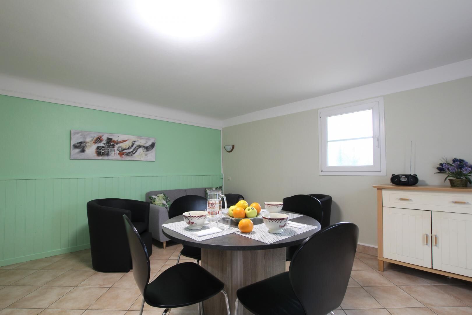 Un coin repas avec une table ronde, des chaises noires, un mur vert, un tableau, une fenêtre et un meuble contenant une radio.