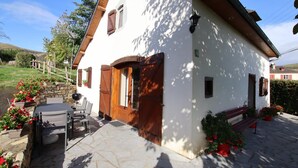 A white building with a stone patio, wooden shutters, and a small outdoor seating area with chairs and a table. - SALATUA - MUSCULDY - Gîte (MUSCULDY)