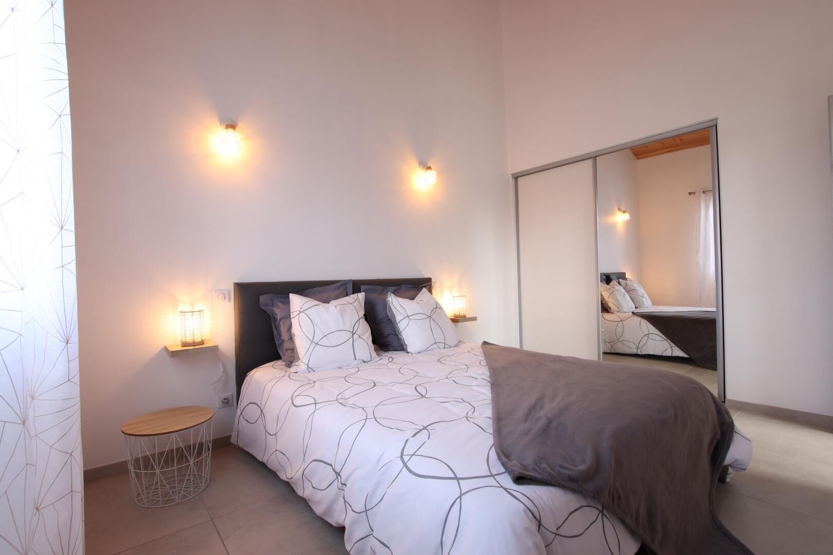 A bedroom with a bed, bedside tables, wall-mounted lights, a mirror, and a geometric-patterned curtain.