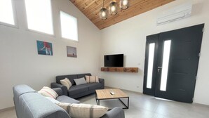 A modern living room with a grey sofa, a coffee table, and a flat-screen TV mounted on a wooden shelf. There are two windows and a door with a glass panel. - JOANESENEA BI - SOURAIDE (SOURAIDE)