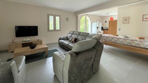 Een moderne woonkamer met een grijze bank, een flatscreen televisie, een houten salontafel en een eethoek met een tafelkleed met een patroon.