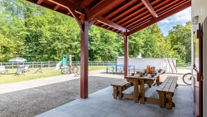 Een overdekt buitengebied met picknicktafels en een speeltuin op de achtergrond.
