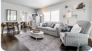 A spacious living room with a grey sofa, a striped armchair, and a round coffee table. There is a dining area with a wooden table and chairs, and a kitchen in the background. - Condo With Heated Pool! Beach & Ocean Access! (Pompano Beach)
