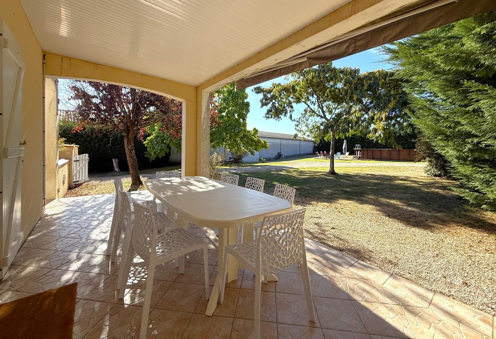 Una veranda coperta con tavolo e sedie, pavimento piastrellato e vista su un giardino recintato con alberi.