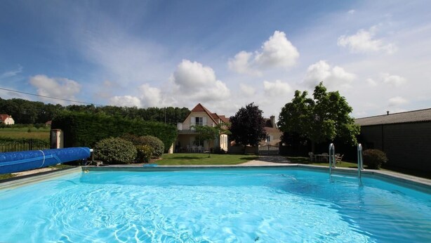 Ein rechteckiger Pool mit einer hölzernen Liegefläche davor, umgeben von Grünpflanzen und einem Haus im Hintergrund.