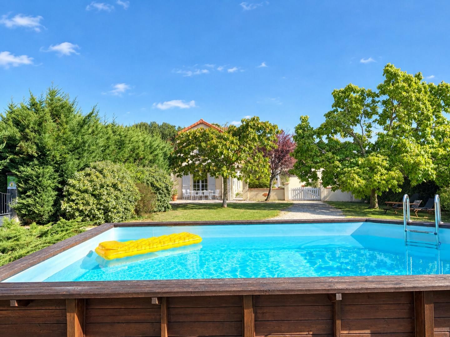 Una piscina con terrazza in legno fronteggiata da alberi e una casa dal tetto rosso.