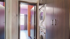 A laundry room with two washing machines and a dryer, a wooden door, and a view into a hallway with a purple wall and a window.