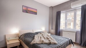 A bedroom with a bed, bedside tables, a nightstand, a window with curtains, and a cityscape view.