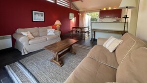 En stue med sofa, sofabord i træ og et tæppe med mønster. Der er et spisebord med stole, og et køkken er synligt gennem en åben dør.