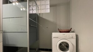 Een praktische wasruimte met een douchecabine van glas, een witte wasmachine en een klein mandje erbovenop.