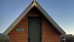 Een houten hutje met de tekst 'BEAR CAVE', een groene deur, houten traptreden en een lantaarn.
