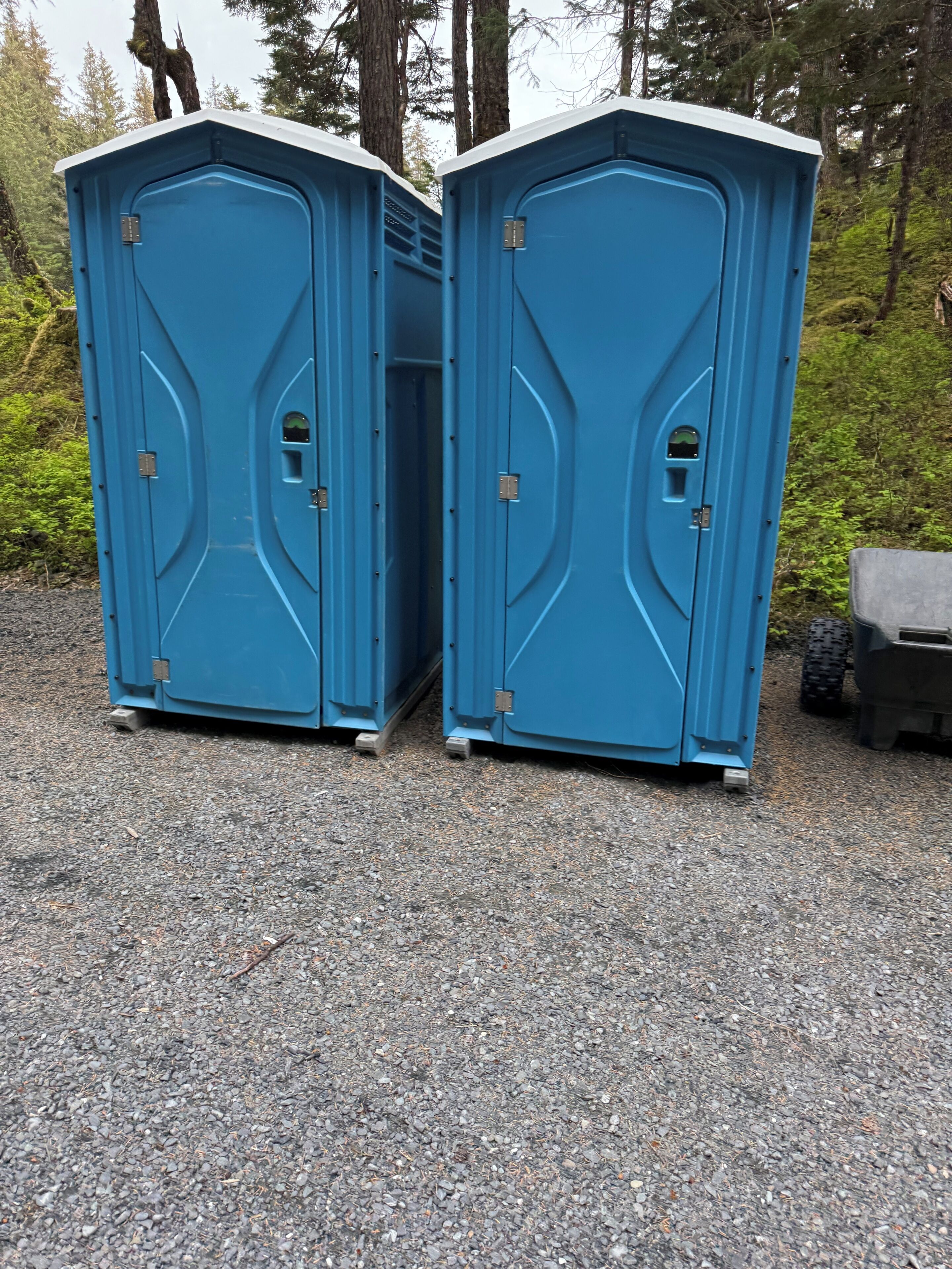 Two portable toilets with a simple design, placed on a gravel surface with trees in the background.