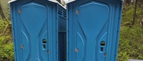 Two portable toilets with a simple design, placed on a gravel surface with trees in the background.