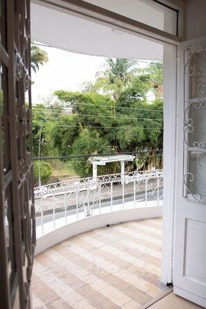 Ein Balkon mit weißer Reling, einer weißen Tür und Blick auf Grünpflanzen sowie ein weißes Gebäude im Hintergrund.