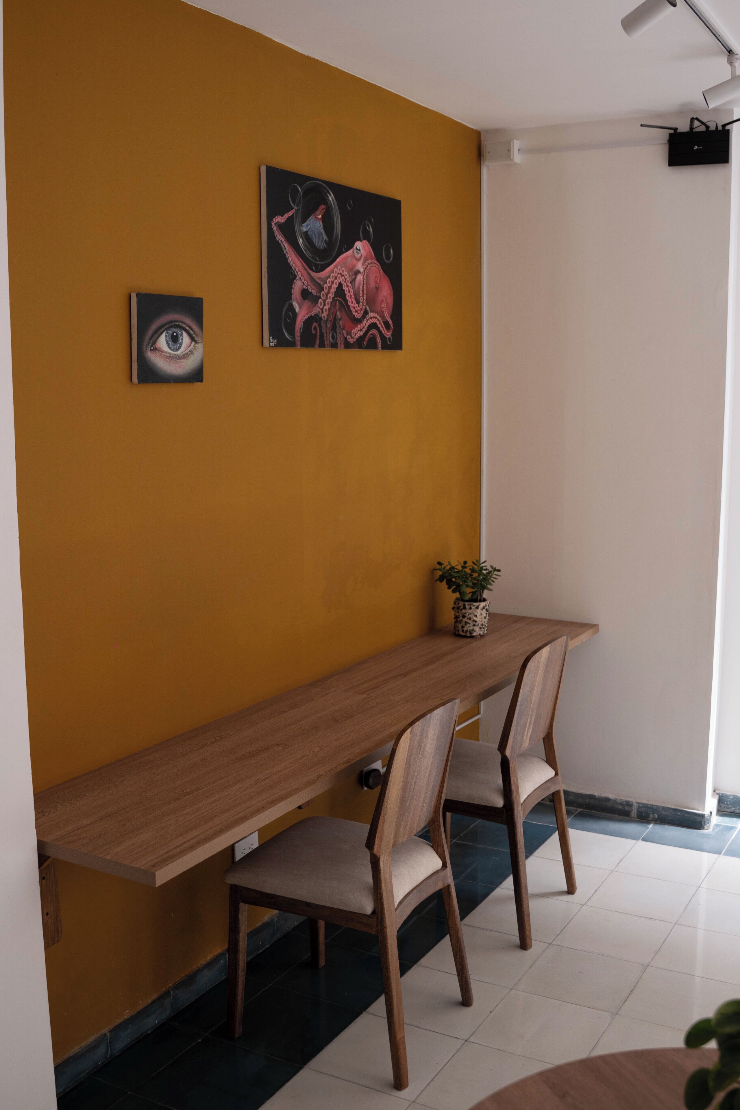 A dining area with a wooden table, two chairs, a potted plant, and two artworks on the wall.