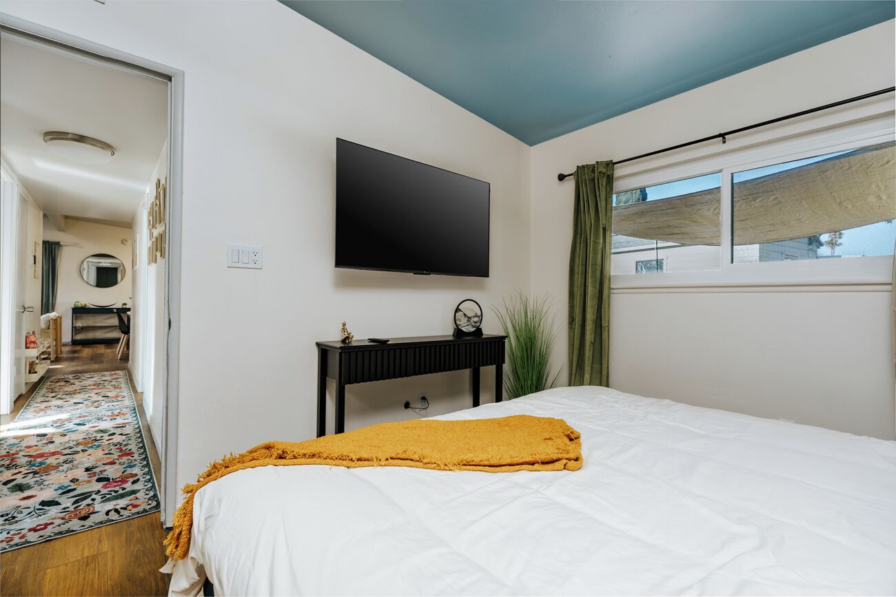 A bedroom with a bed, a TV, a bedside table, a window with curtains, and a hallway leading to another room.
