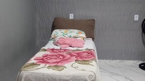 A small, neatly made bed with a floral bedspread and a pink pillow in a room with grey walls and a tiled floor. - Ksl bird refuge (Araquari)