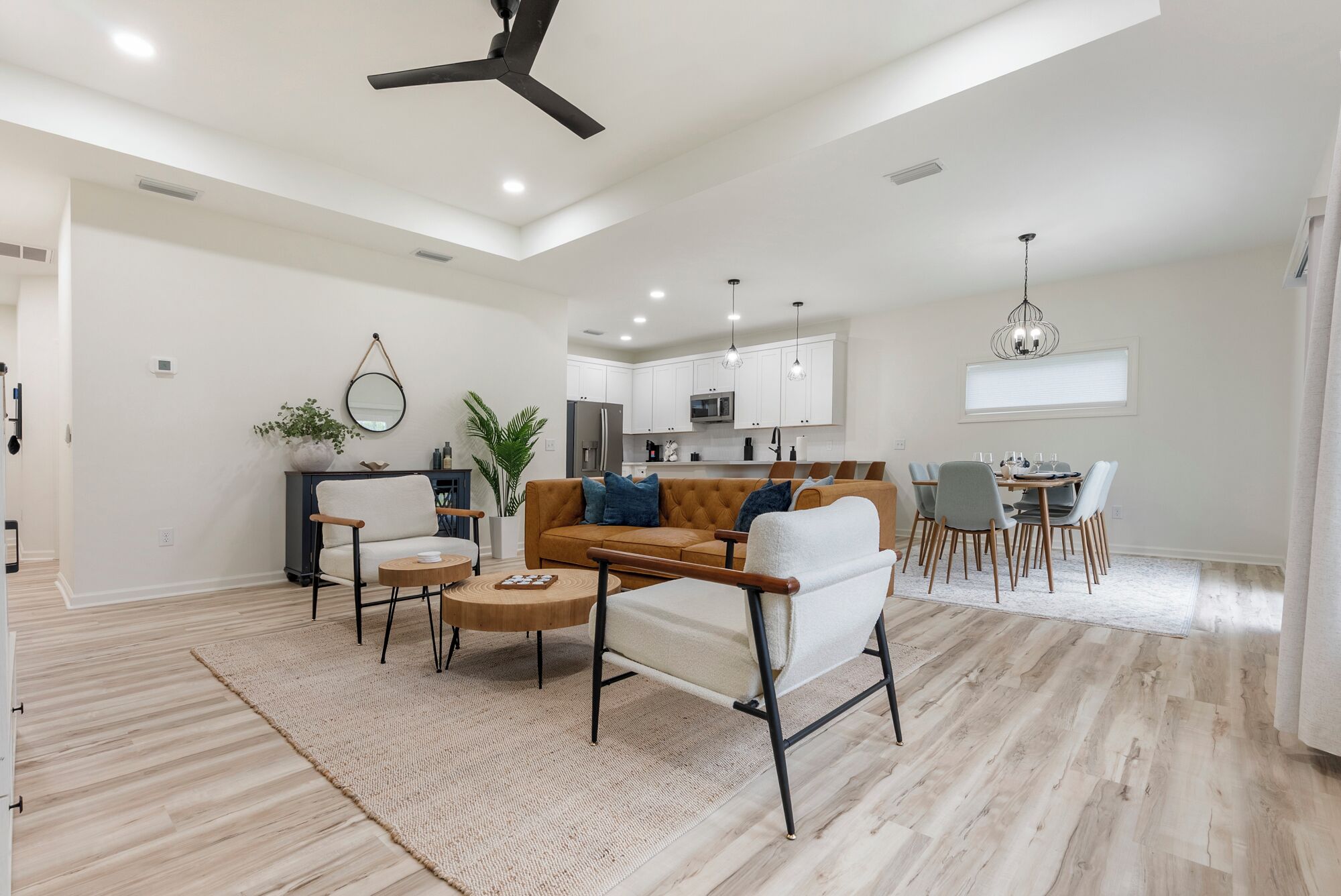 Un salon spacieux avec un canapé, des fauteuils et une table basse. Au fond, on aperçoit une cuisine avec des meubles blancs et un coin repas comprenant une table et des chaises.