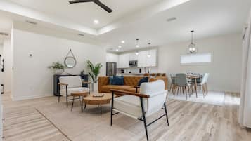 Un salon spacieux avec un canapé, des fauteuils et une table basse. Au fond, on aperçoit une cuisine avec des meubles blancs et un coin repas comprenant une table et des chaises.