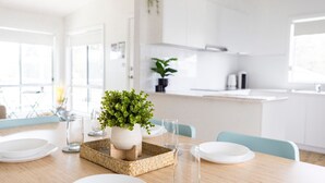 A modern kitchen with white cabinets, a central island with a plant, and a dining table set with plates and glasses. - Stay and Float in Basin Bliss (Saint Georges Basin)