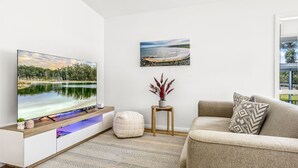 A modern living room with a flat-screen TV, a wooden entertainment unit, a beige sofa, a patterned cushion, a small wooden side table with a vase, and a framed landscape painting on the wall. - Stay and Float in Basin Bliss (Saint Georges Basin)