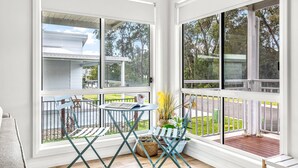 A room with a view of a white building and a grassy area. There is a small table with a chair and a potted plant. - Stay and Float in Basin Bliss (Saint Georges Basin)