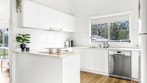 A modern kitchen with white cabinetry, a built-in dishwasher, and a window with a view of trees. - Stay and Float in Basin Bliss (Saint Georges Basin)