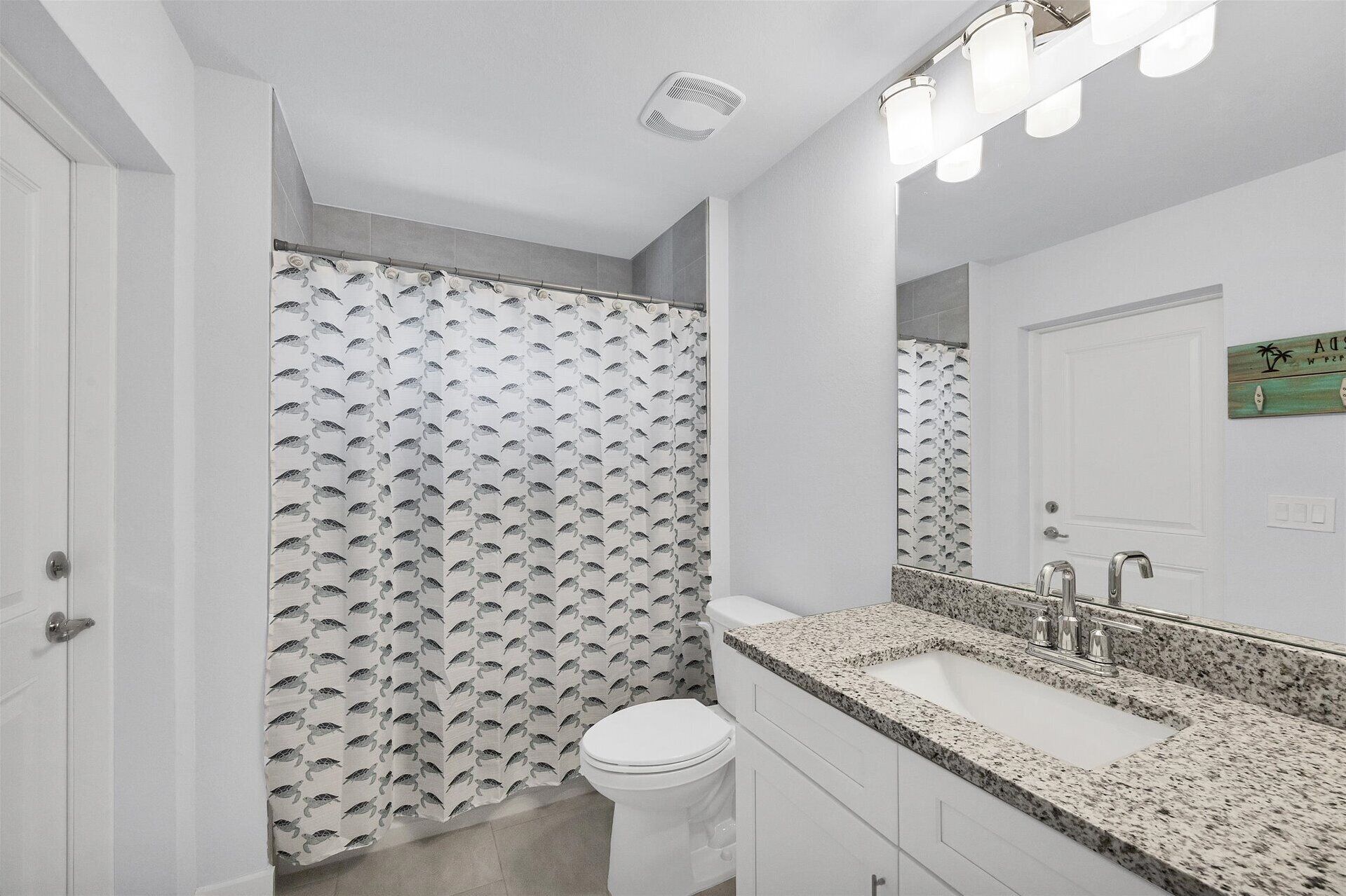 A bathroom with a shower curtain featuring a shark pattern, a white toilet, a granite countertop sink with a modern faucet, and a mirror with lights above it.