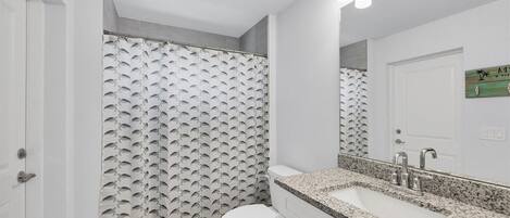 A bathroom with a shower curtain featuring a shark pattern, a white toilet, a granite countertop sink with a modern faucet, and a mirror with lights above it.