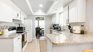 Una cocina con gabinetes blancos, encimera de granito, comedor con mesa y sillas, y ventilador de techo.