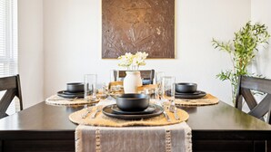 Una mesa de comedor dispuesta con cuencos, platos y cubiertos negros, un florero blanco con flores y un cuadro abstracto grande en la pared.